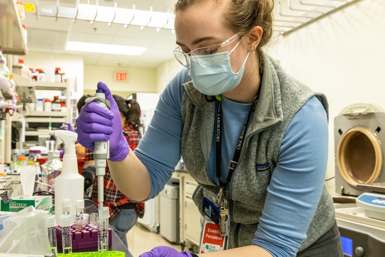 A scientist in a lab coat and mask using a pipette at a laboratory bench