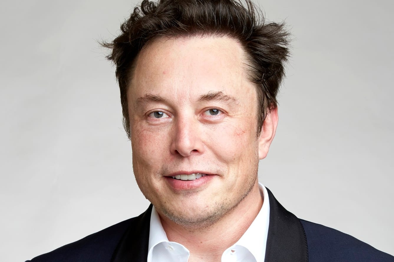 Elon Musk smiling, wearing a dark jacket and an open white shirt