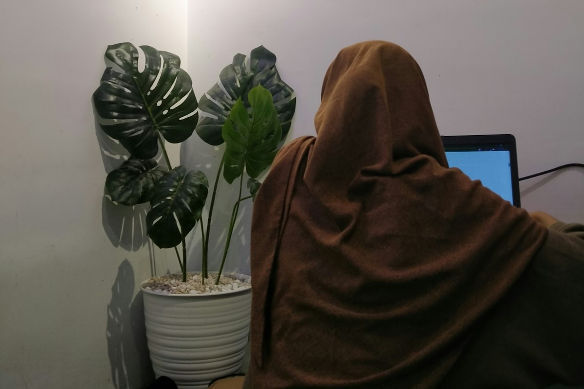 A woman in a headscarf, seen from behind, working on a laptop beside a potted plant
