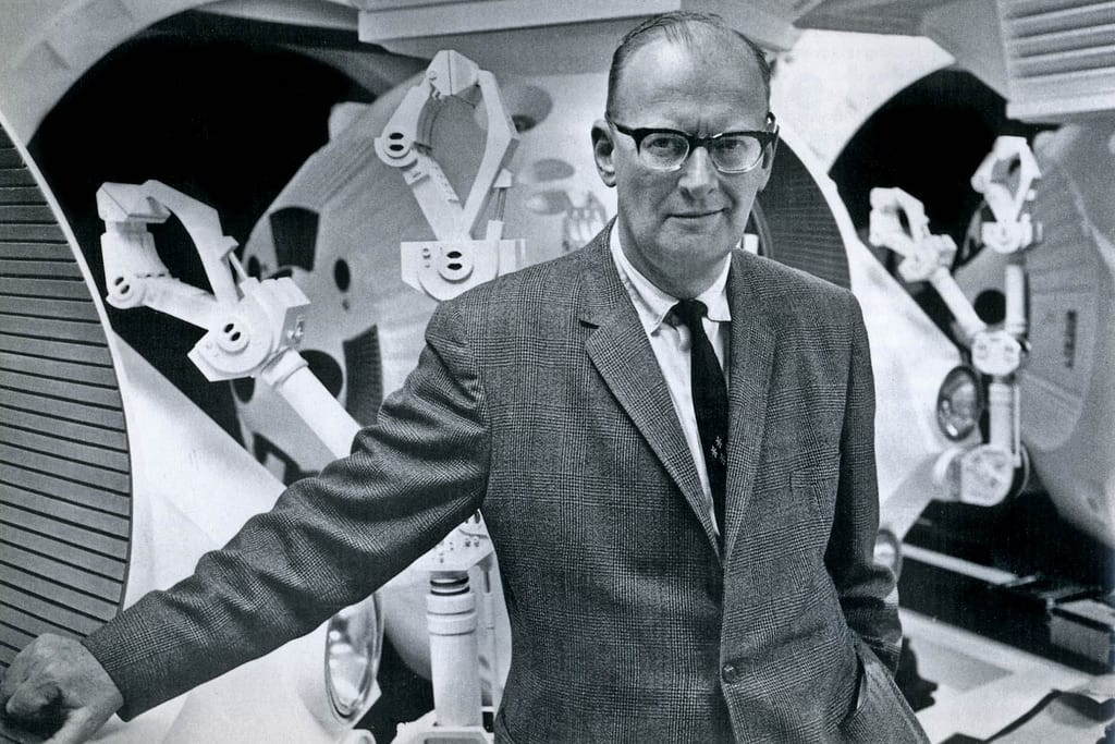 Arthur C. Clarke in glasses and a suit, standing between large robotic arms