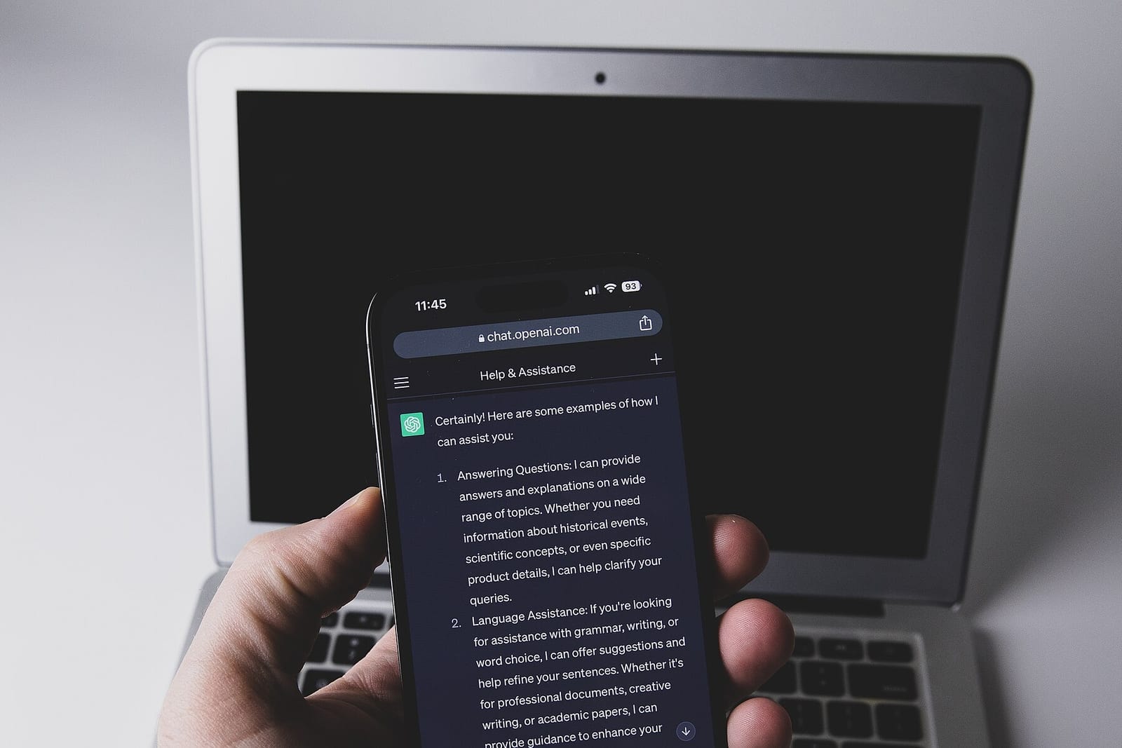 A hand holding a smartphone with the ChatGPT app open in front of a laptop