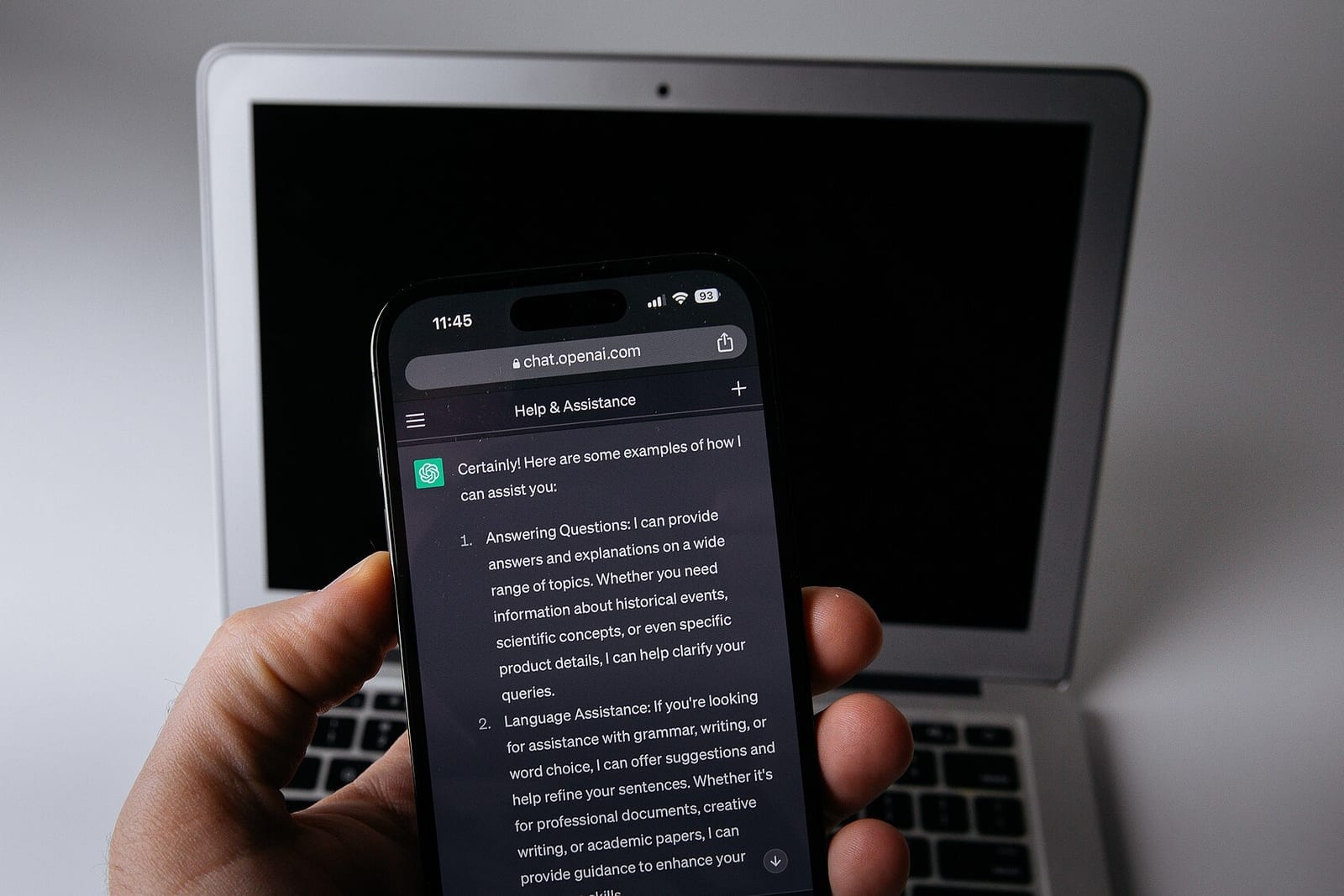 A hand holding a smartphone with ChatGPT open in front of a laptop